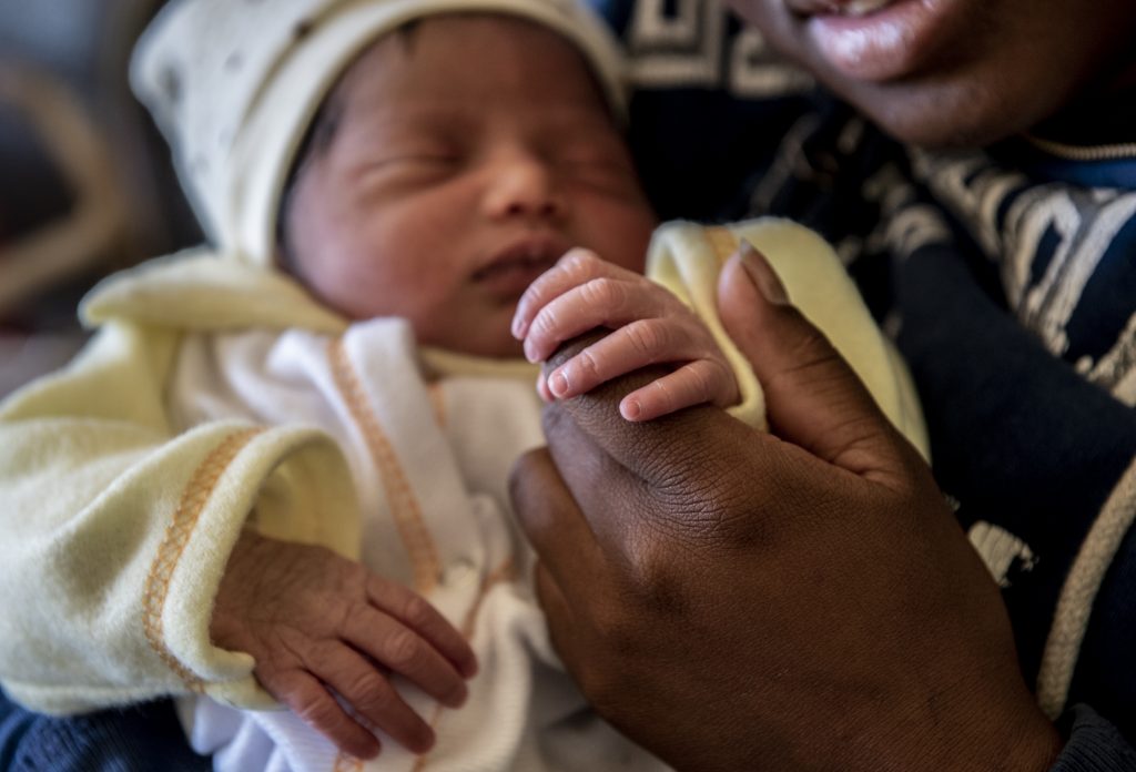 Newborn girl wearing a sweater and cap being held in a woman's arms.