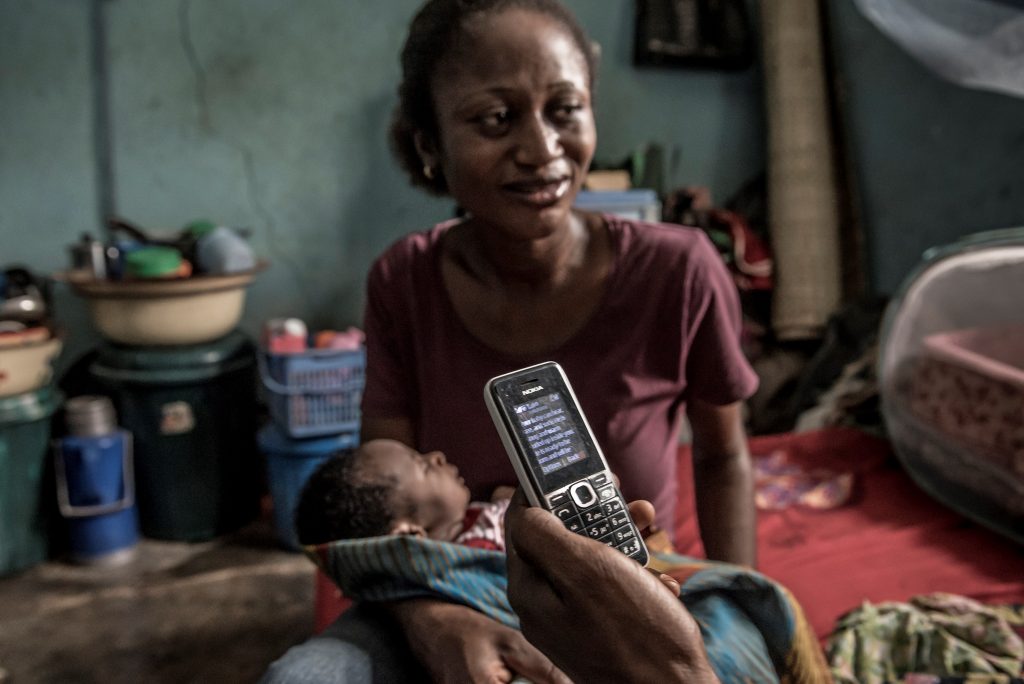 A woman holds her baby as someone holds a cellphone with text.