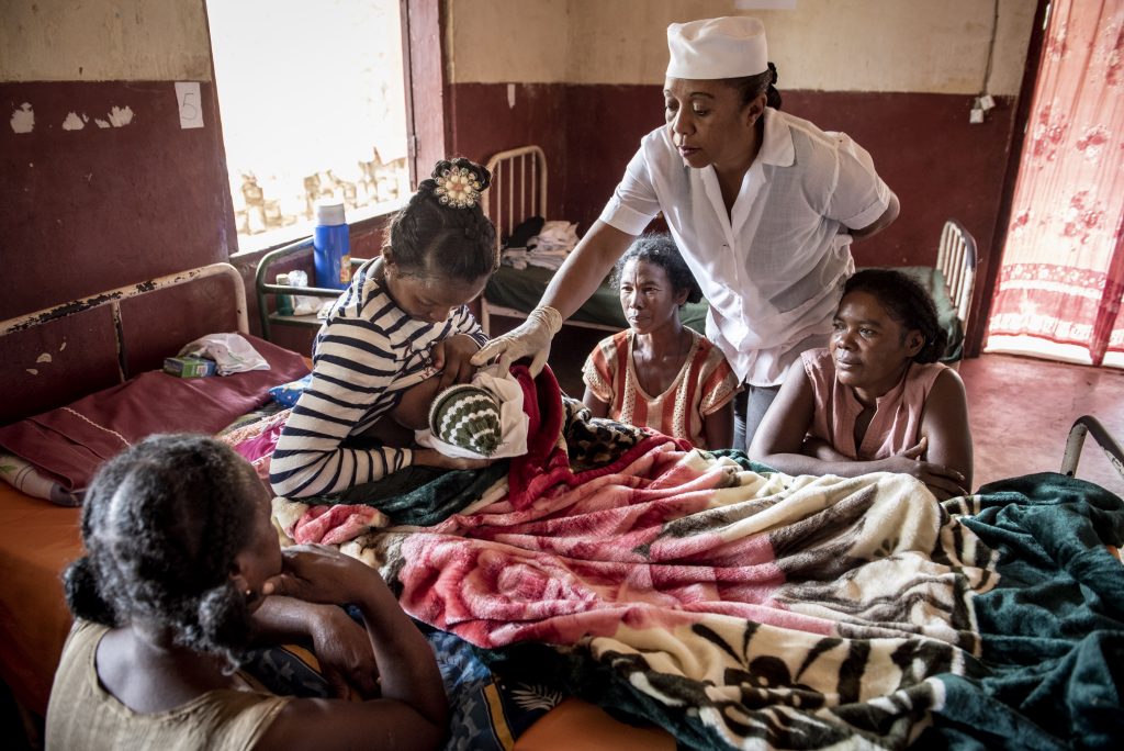 Three women gather around a new mother and baby as a nurse gives her advice about breastfeeding in a health facility.
